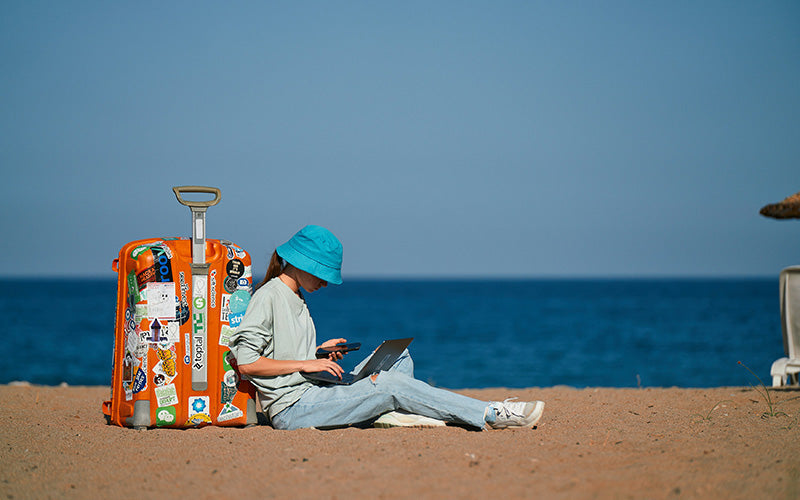 Women sitting on beach leaning against suitcase using laptop
